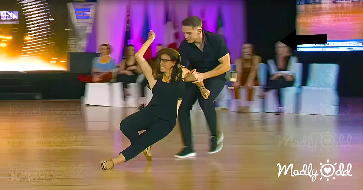 A couple dances gracefully on a wooden floor, the woman in a black outfit and gold shoes, the man in black attire.