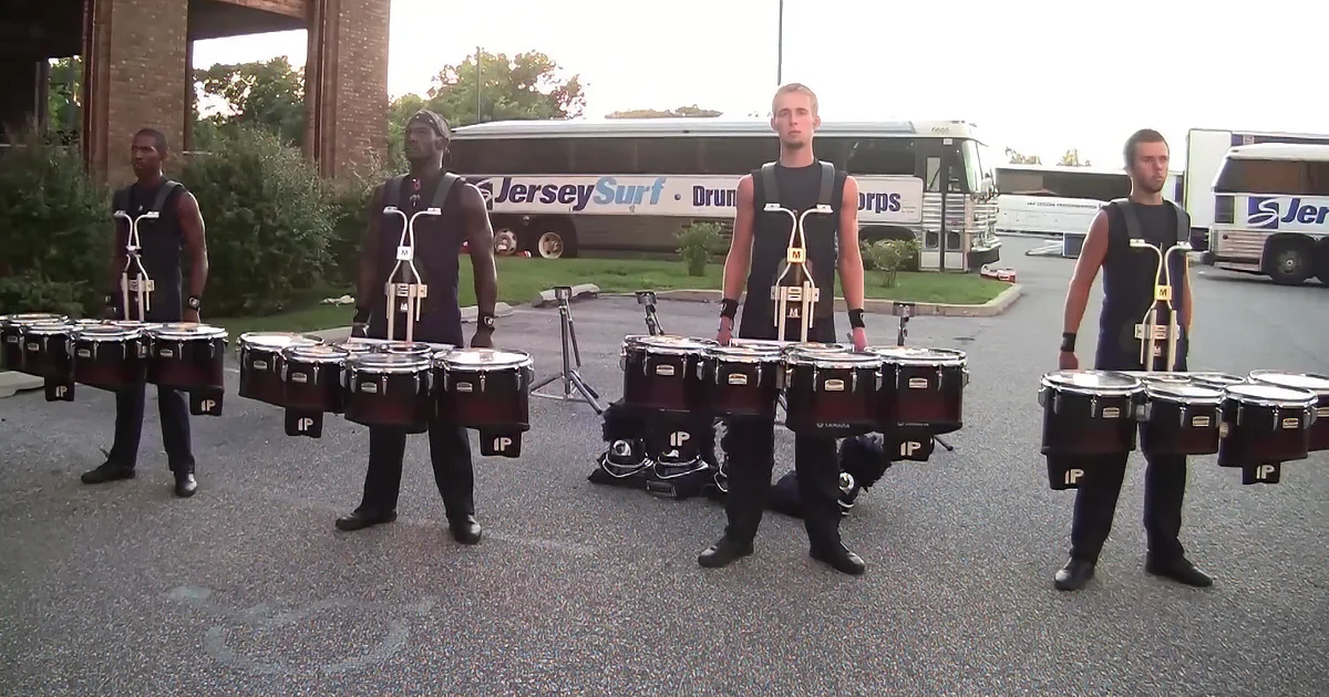 Four drummers in black outfits play timpani in a parking lot near a "Jersey Surf Drum & Bugle Corps" bus.