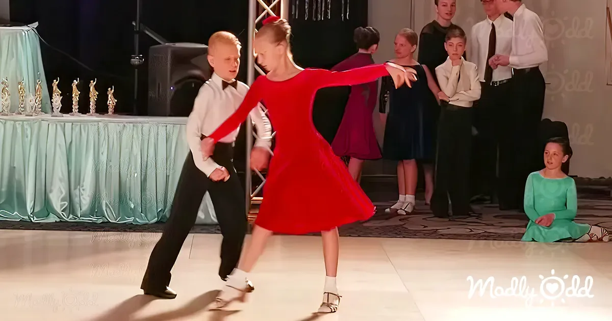 A young girl in a red dress dances gracefully with a boy in a white shirt and black pants, surrounded by spectators.
