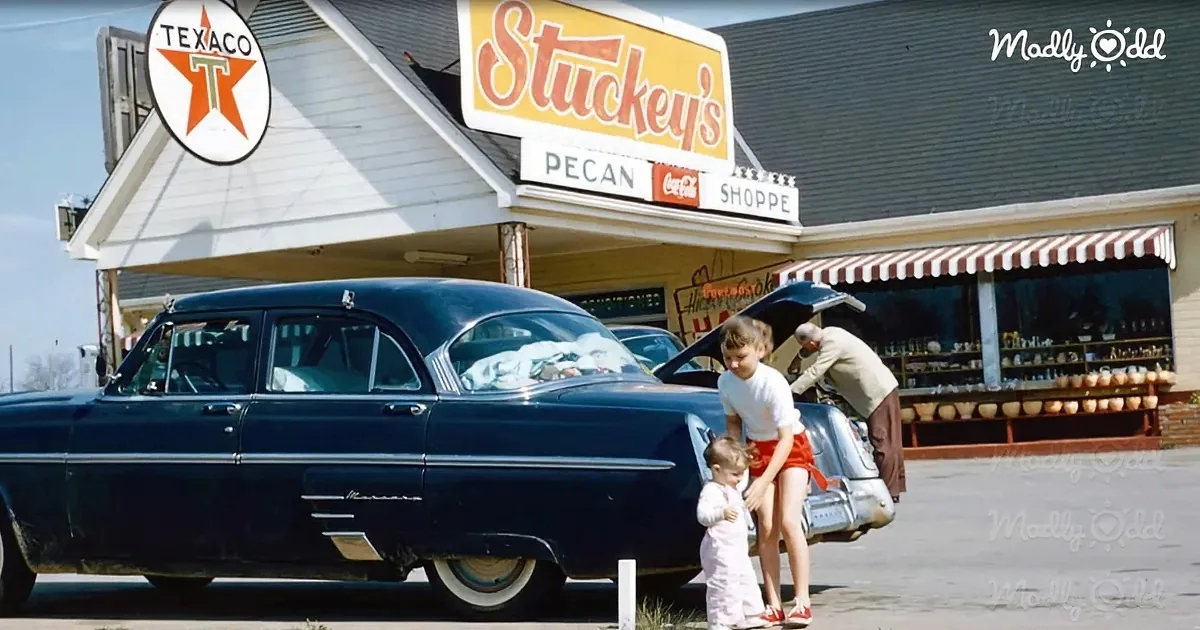 A vintage blue car is parked in front of a Stuckey's Pecan Shoppe with people interacting near the trunk.