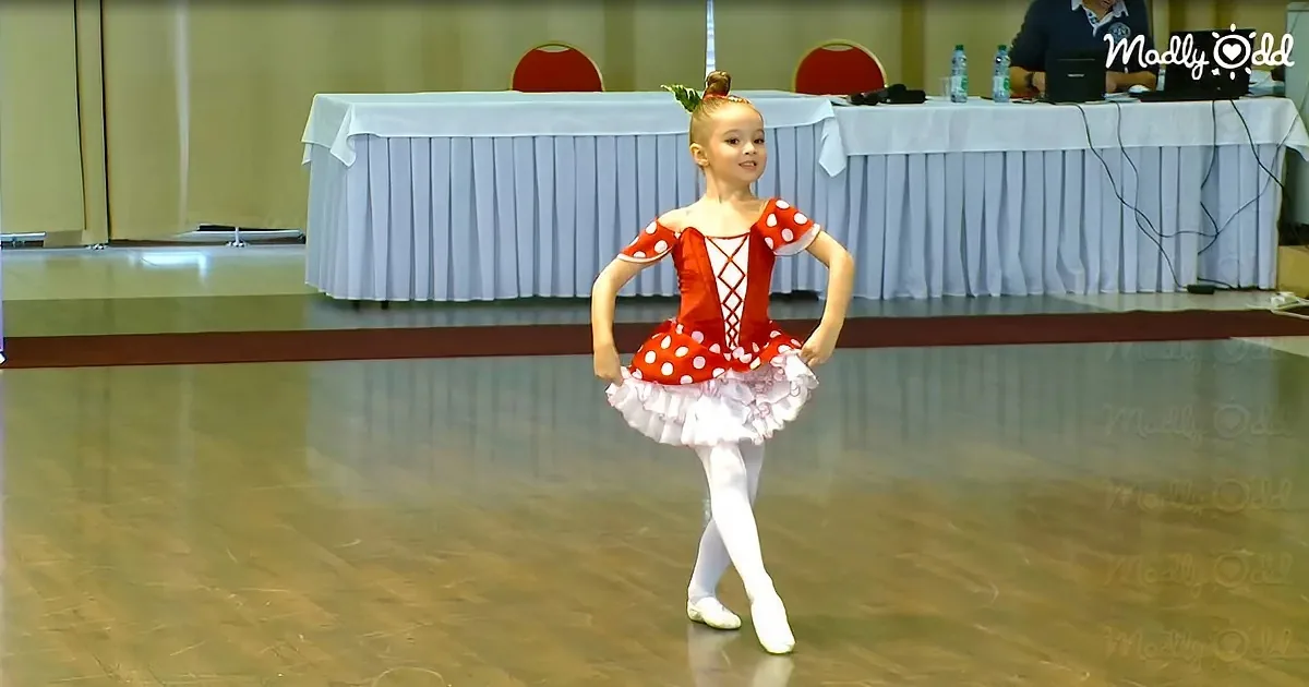 A young girl in a red polka-dot ballet costume stands on a stage with her hands on her hips.
