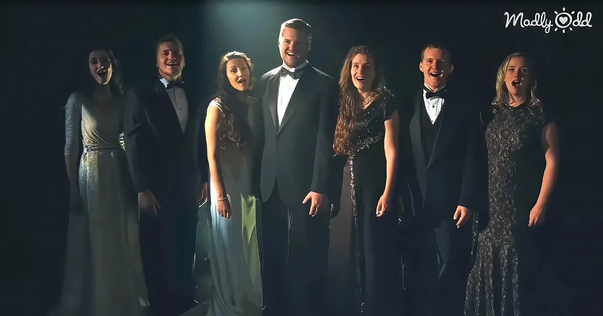 A group of six people in formal attire stands together, singing under a spotlight.