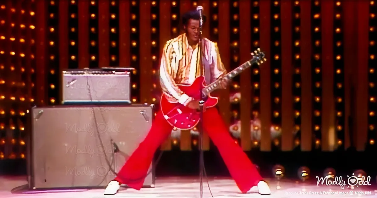 A musician in a striped shirt and red pants plays a red guitar on stage with amplifiers and a backdrop of lights.
