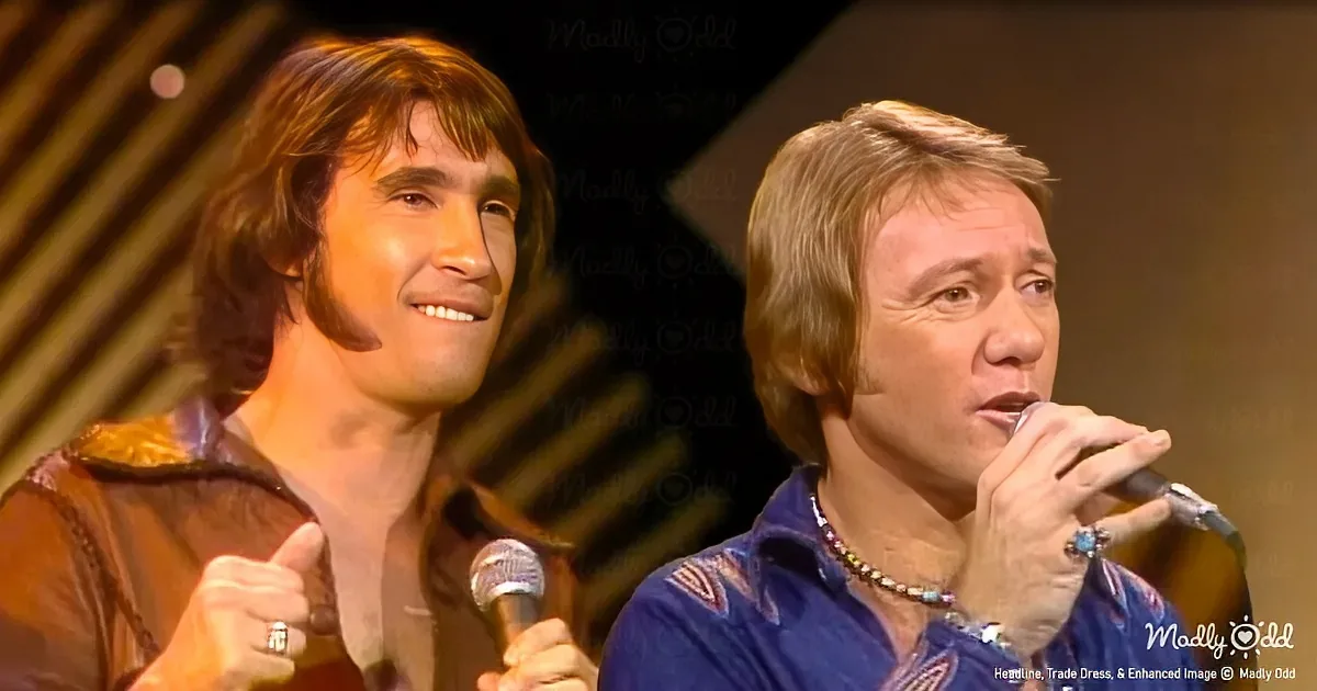 Two men on stage, one singing into a microphone, the other giving a thumbs-up gesture.
