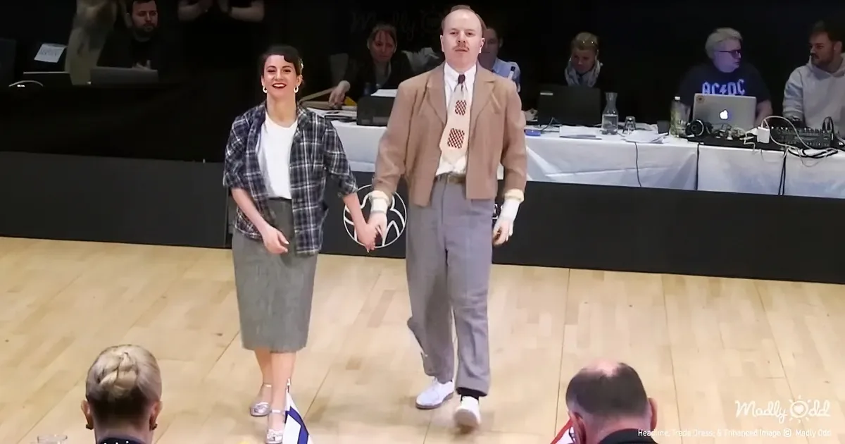 A man and woman walk hand-in-hand on a stage, both dressed formally, with an audience and panelists in the background.