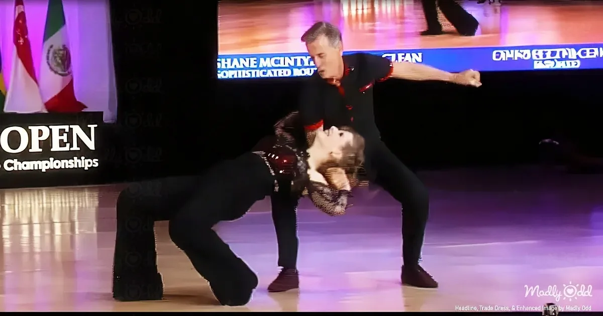 A man and woman are dancing on a stage, the man in a black outfit and the woman in a red and black dress.
