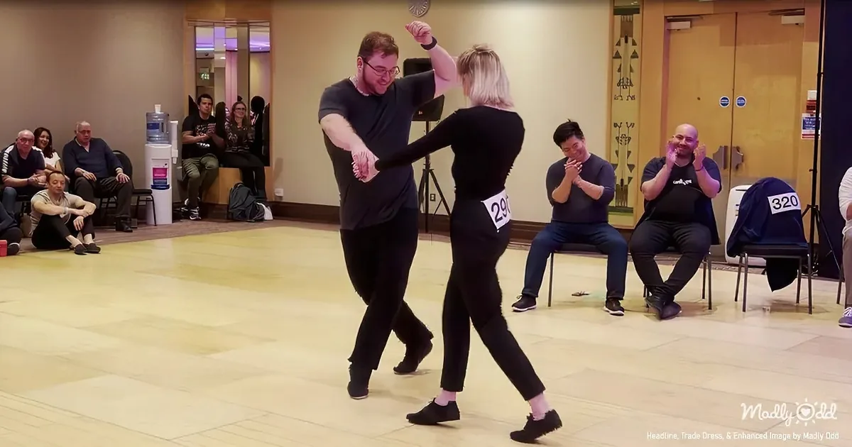 A man and woman dance together in a room with spectators watching and clapping.