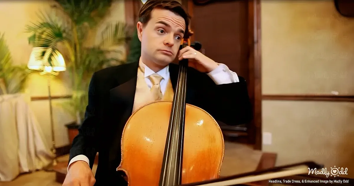 A man in a suit plays a cello in a warmly lit room with plants and a lamp.