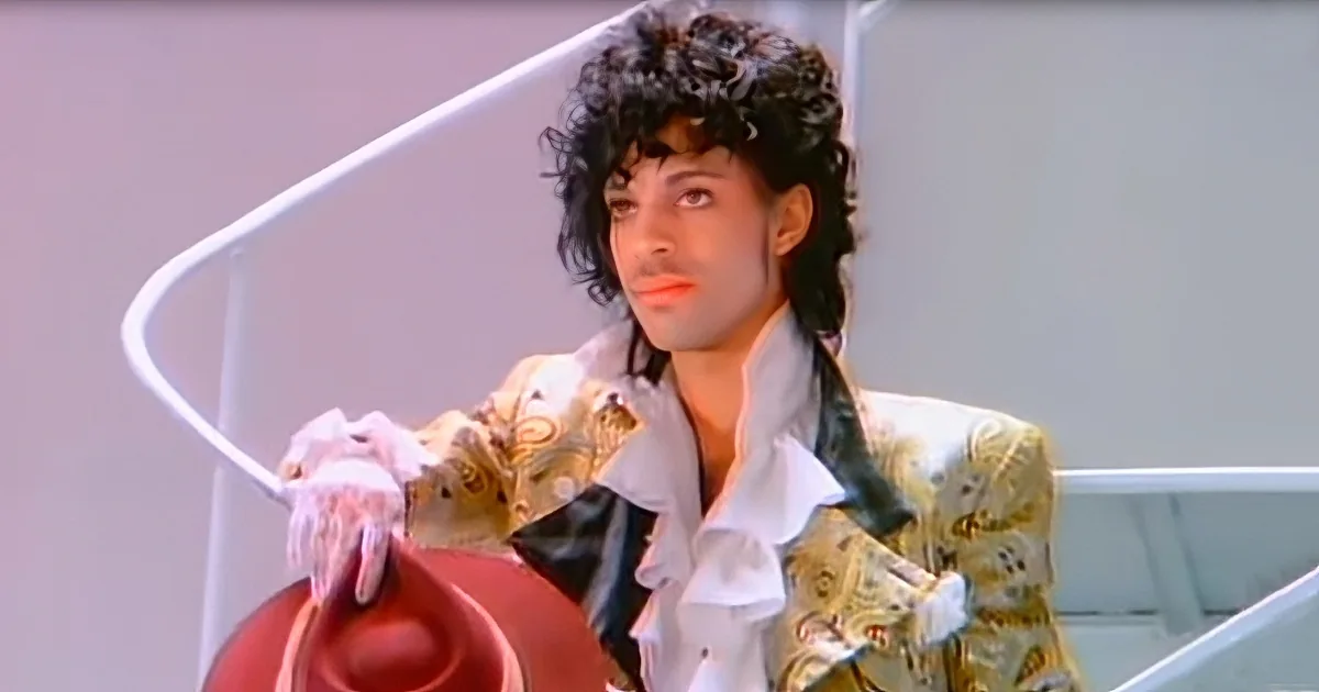 Prince in a white shirt and patterned jacket, holding a red hat, with curly hair and makeup, leaning on a white railing.