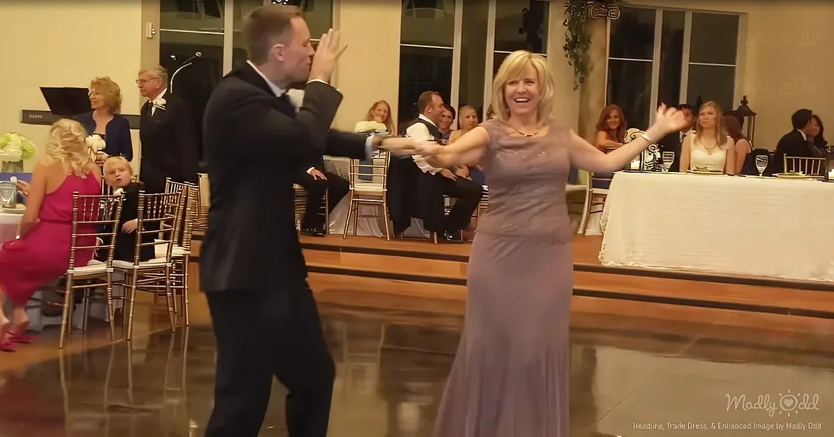 A man and woman in formal attire dance joyfully on a polished floor, surrounded by seated guests in a warmly lit room.