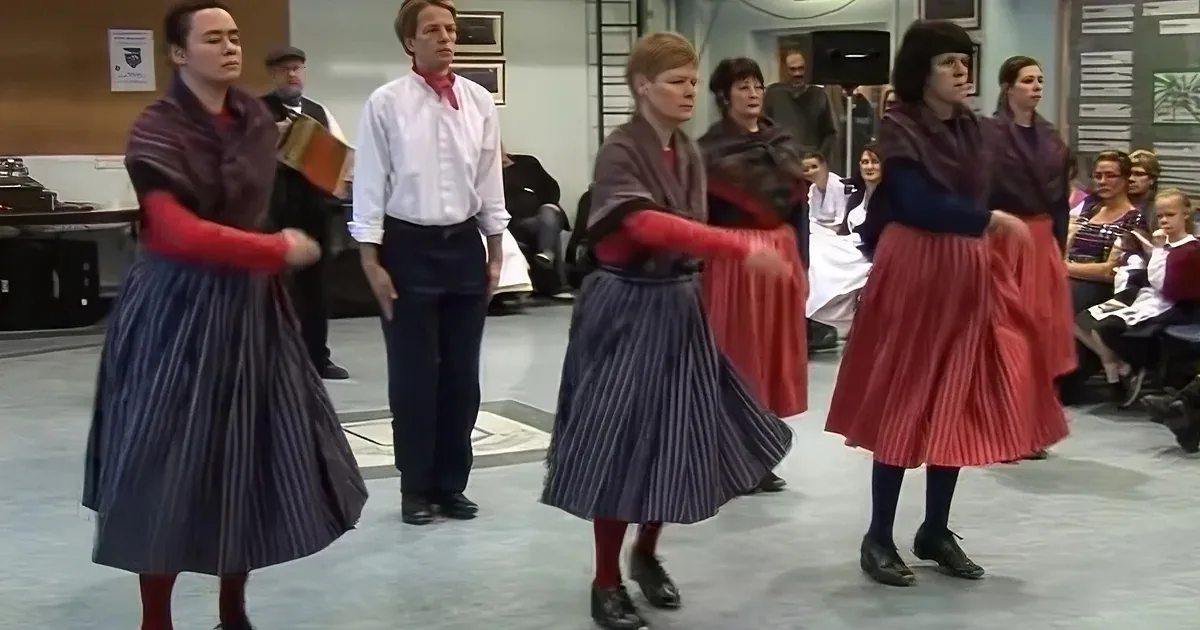 Women in pleated skirts and red tights are dancing, with one holding a book, while spectators watch.