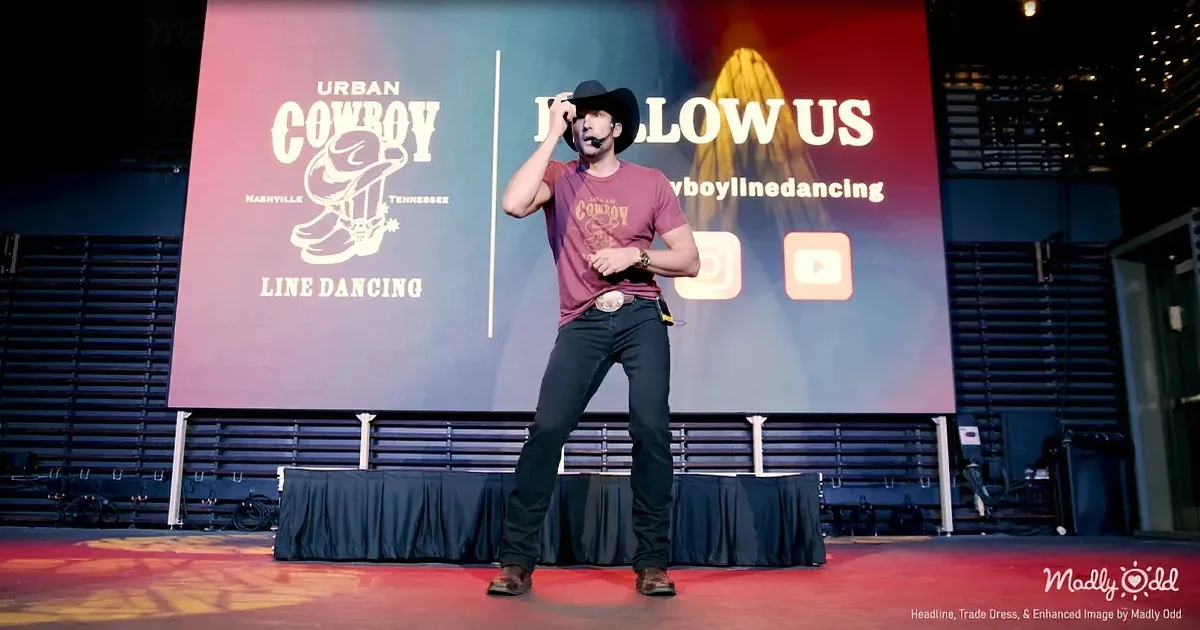 A man in cowboy attire stands on stage, holding a microphone, with a backdrop promoting "Urban Cowboy Line Dancing."