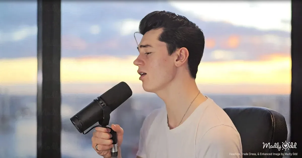 A person sings into a microphone, wearing a white shirt, against a sunset backdrop.