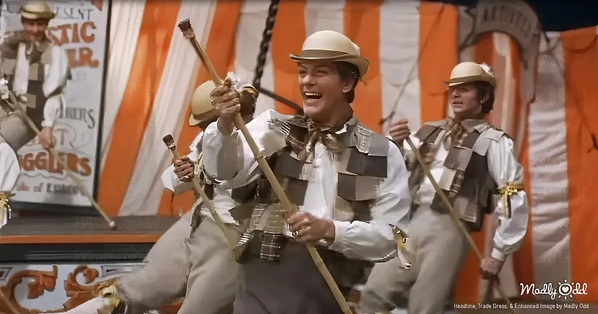 A man in a white shirt and hat, holding a stick, smiles while performing in a circus-like setting.