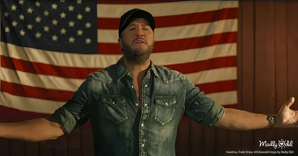A man in a denim shirt and cap stands with arms outstretched in front of an American flag.