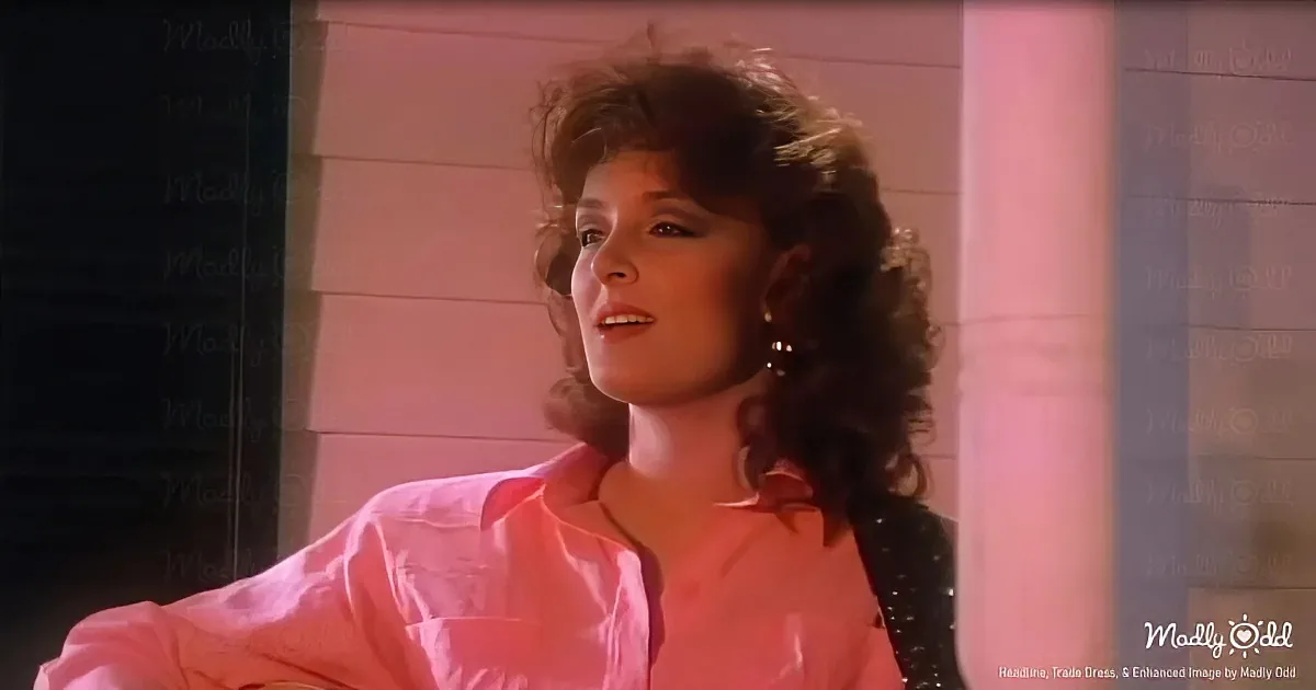 A woman with curly hair, wearing a pink shirt, stands indoors with a light pink hue.