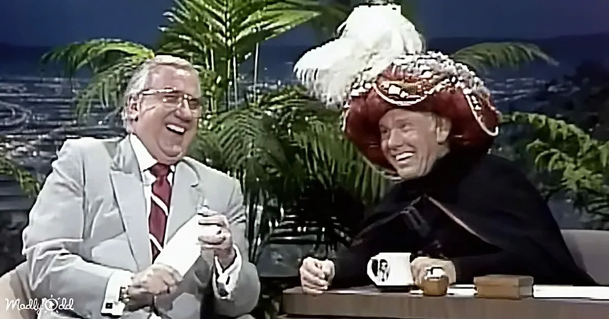 Two men in suits laugh, one in a red hat and black outfit, another in a light suit and red tie.