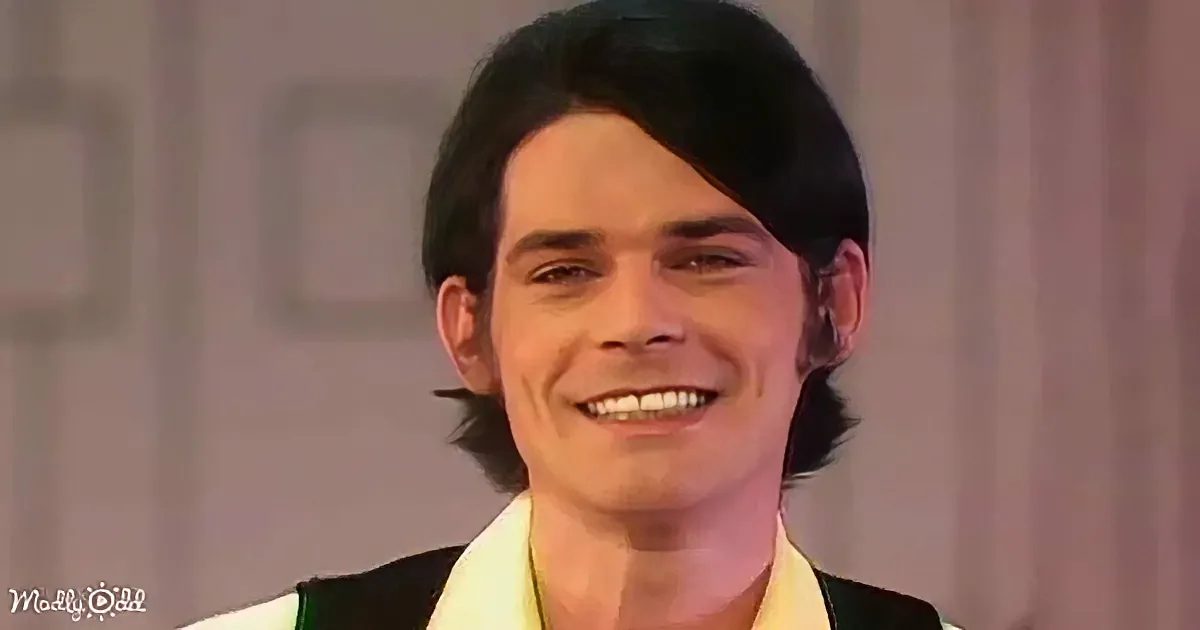 A smiling person with dark hair, wearing a white and black outfit, against a blurred background.