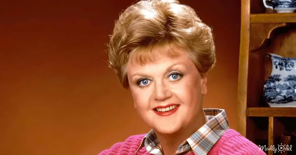 A smiling woman with short, styled hair, wearing a pink sweater over a plaid shirt, stands in front of a wooden shelf...