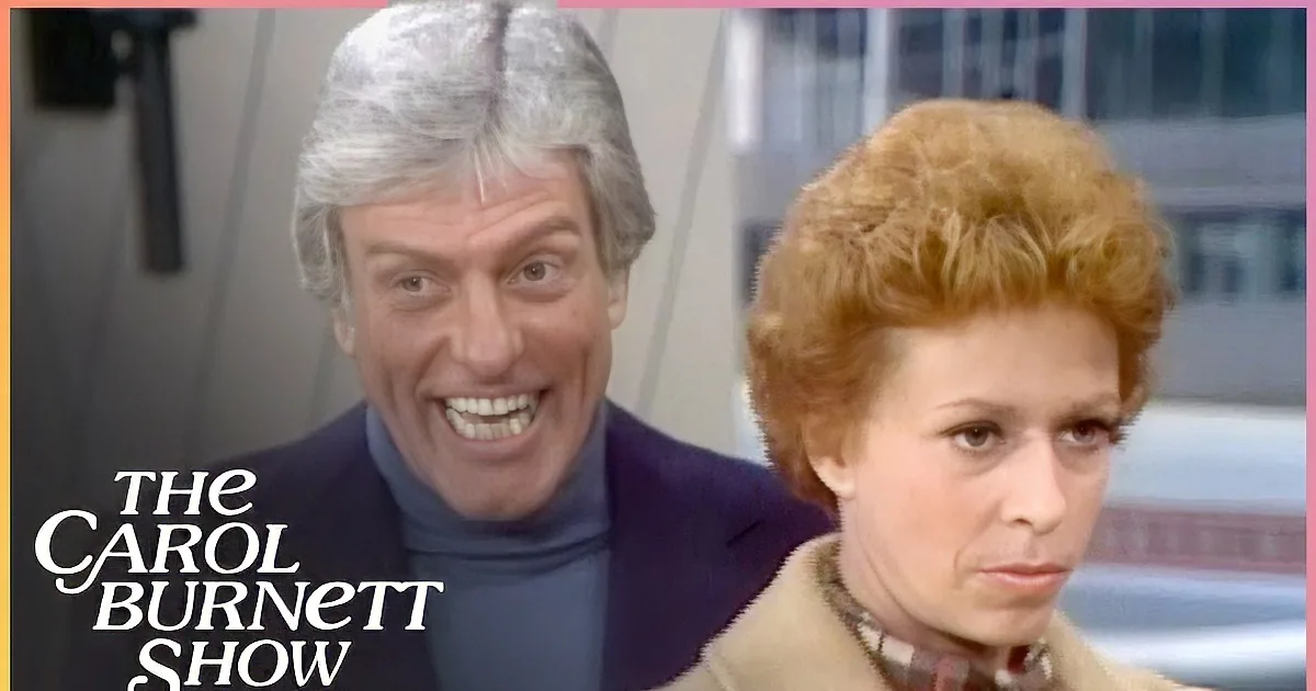 A man in a blue suit points while a woman in a red plaid shirt looks on, with others in the background.