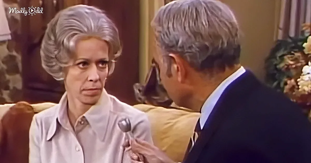 A woman with a large, styled wig sits on a couch, looking at a man in a suit who is holding a spoon.