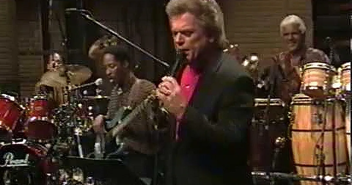 A man in a suit sings passionately, surrounded by musicians and instruments.