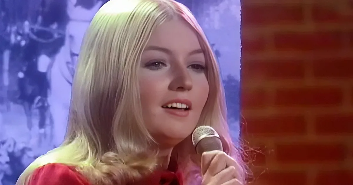 A woman with blonde hair, wearing a red outfit, holds a microphone in front of a brick wall and a blue background.