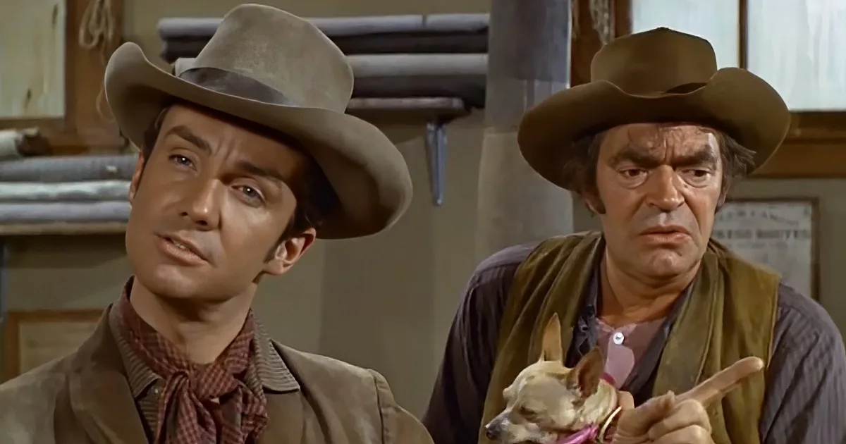 Two men in cowboy hats, one holding a small dog, appear to be in a Western setting.