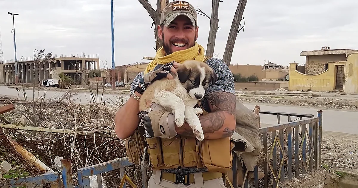 A man in military gear holds a puppy, smiling, amidst a backdrop of damaged buildings and debris.
