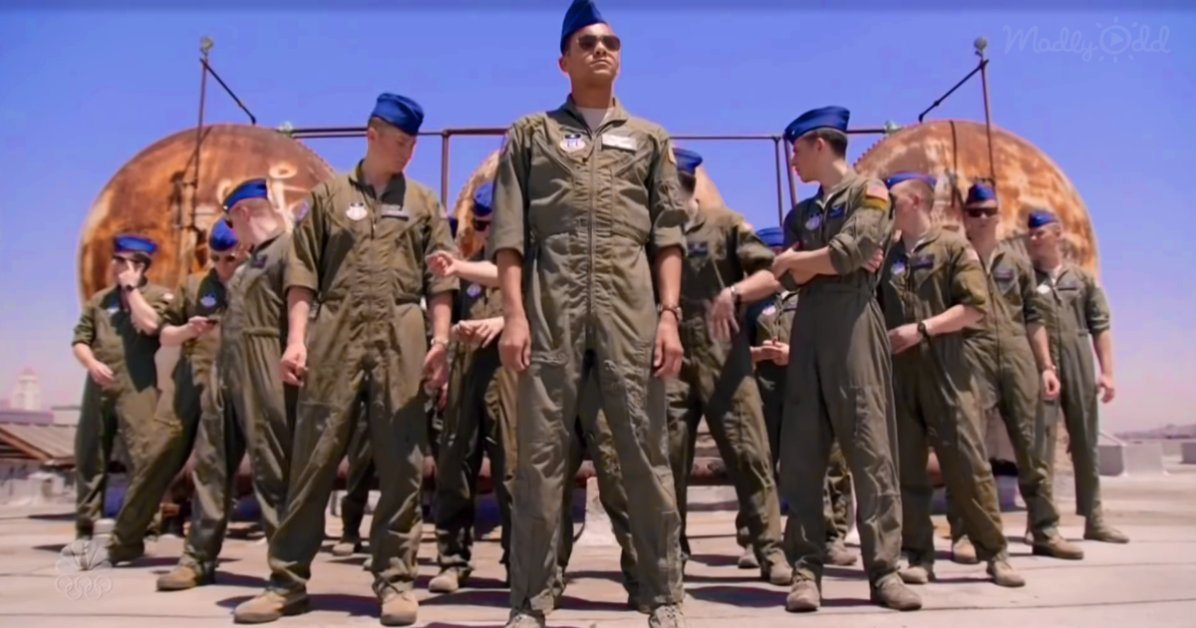 A man in a flight suit stands in the center, surrounded by uniformed personnel, with a large rusted structure in the...