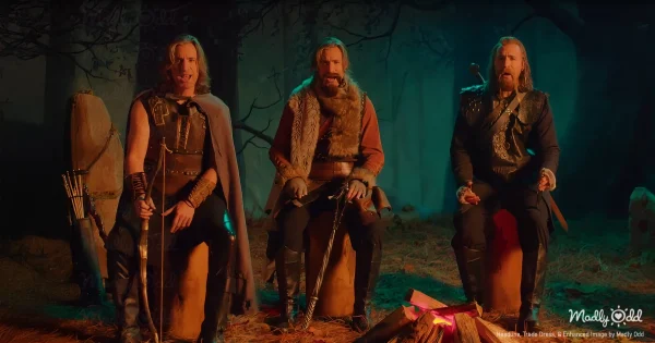 Geoff Castellucci stands dressed as four distinct dwarves, each holding their unique weapon. Behind them, a mountain smolders in the background, with smoke billowing into a dark, fiery sky.