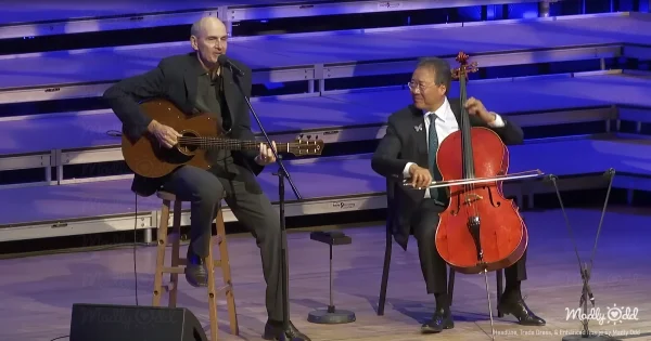 Yo-Yo Ma standing with his cello under the soft lights of a Tanglewood stage, James Taylor beside him with his guitar. Both legends locked in a deep musical connection, performing with an intimate crowd in awe.