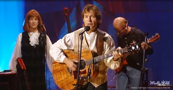 John Denver stands on stage under dim, moody lighting, strumming an acoustic guitar as the spotlight focuses on him. Behind him, James Burton plays electric guitar, his presence understated but powerful, while the audience sits captivated.