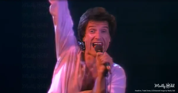 A vibrant, 1980s concert stage lit with intense white and blue spotlights, Ray Davies belting out lyrics in front of a massive crowd while Dave Davies plays his iconic guitar solo. The audience is on their feet, hands raised, completely engrossed in the moment.