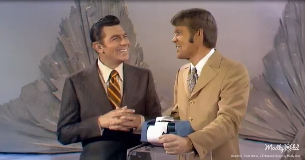 Glen Campbell and Andy Griffith stand together on a 1960s television variety show set with festive decorations. The scene captures warm stage lighting, vintage broadcast cameras, and classic Christmas decorations typical of 1969 television productions.