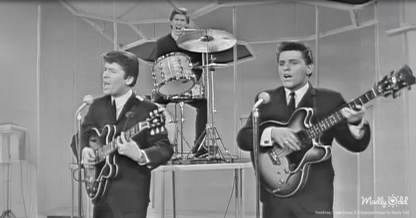 A black-and-white 1960s TV stage with The Searchers sharply dressed in suits, playing guitars with total focus. The audience, a mix of young and old, leans forward, eyes fixed on the stage as if they’re witnessing magic.